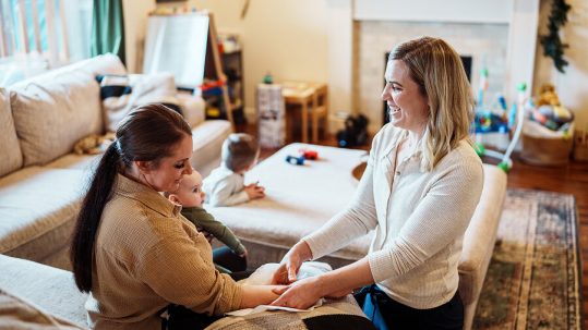 A smiling woman sitting in a home setting with a baby on her lap. Another woman is smiling and attending to her arm.