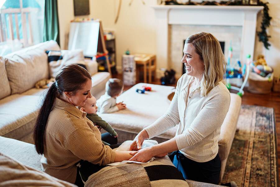 A smiling woman sitting in a home setting with a baby on her lap. Another woman is smiling and attending to her arm.